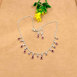 Op de afbeelding: Een zilverkleurige ketting en bijpassende oorbellen met kleine, druppelvormige rode stenen die aan een fijne ketting hangen. De ketting heeft meerdere stenen accenten over de lengte en de oorbellen weerspiegelen het druppelontwerp.