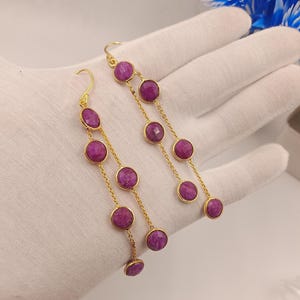 Op de afbeelding: Een paar goudkleurige oorhangers met ronde, dieproze edelstenen. De edelstenen zijn gezet in goudkleurige bezels en verbonden door fijne gouden kettingen. De oorbellen worden gepresenteerd tegen een witte achtergrond.