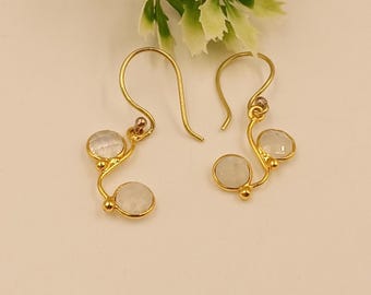 May include: A pair of gold-colored earrings with a curved design. Each earring features two round, light-colored stones. The earrings have a hook closure and are displayed on a white surface with a green and white floral accent.