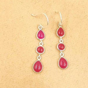 May include: A pair of drop earrings featuring silver-toned hooks and three vertically aligned, smooth, dark pink stones. Each earring consists of a small teardrop-shaped stone at the top, a circular stone in the middle, and a larger teardrop-shaped stone at the base, all connected by small silver-toned metal links.