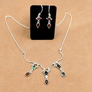 Puede incluir: Un conjunto de collar y pendientes colgantes de plata con piedras preciosas ovaladas de varios colores. El collar tiene una cadena de plata curva con tres secciones colgantes, cada una con dos pequeñas piedras en tonos verdes, marrones y negros.