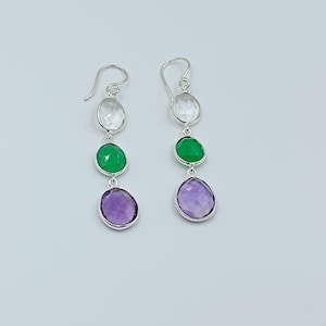 Puede incluir: Un par de pendientes colgantes de plata con tres piedras preciosas de colores. La piedra superior es transparente, la central es verde y la inferior es morada. Las piedras son ovaladas y están engarzadas en plata.
