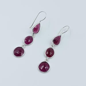 May include: A pair of dangle earrings with a silver-tone metal setting. Each earring features three faceted, oval, and round-cut, deep red gemstones. The earrings hang from a curved hook.