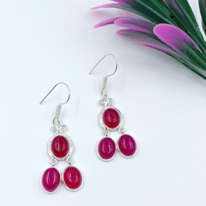 May include: Silver drop earrings featuring three oval, magenta gemstones. The earrings have a decorative silver design above the top stone and a hook fastening. They are presented on a white surface with purple and green floral accents.