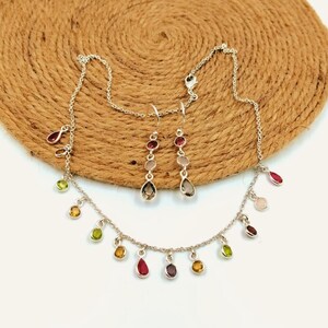 Puede incluir: Un collar de plata y un par de pendientes a juego con pequeños dijes de piedras preciosas multicolores en varias formas, incluyendo gotas y redondas. La cadena del collar está adornada con piedras colgantes en tonos rojos, verdes y amarillos.