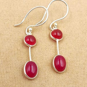 Puede incluir: Pendientes colgantes plateados con piedras rojas. Cada pendiente tiene una pequeña piedra roja redonda sobre una piedra roja ovalada más larga. El fondo es de color marrón claro neutro.
