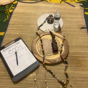 Puede incluir: Un surtido de artículos espirituales que incluyen una cara tallada en madera, una botella de agua, una escultura de madera, un collar de cuentas con conchas y una portapapeles con un bolígrafo. Los artículos están dispuestos sobre una estera tejida.