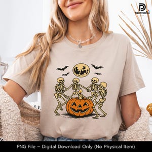 May include: Beige t-shirt featuring a Halloween design. The graphic shows skeletons dancing around a jack-o'-lantern with a candle, a full moon, and bats. The text at the bottom reads: PNG File - Digital Download Only (No Physical Item).
