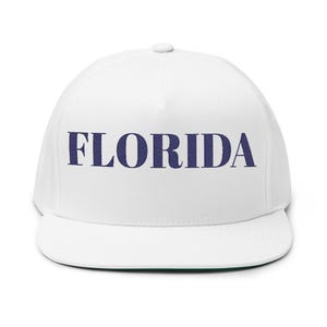 May include: A white flat-brimmed cap featuring the word "FLORIDA" embroidered in navy blue. The cap has a structured crown and a flat peak, with a green underside.