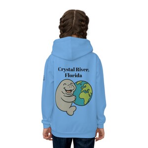 May include: Light blue hoodie featuring a cartoon manatee hugging a globe. The text "Crystal River, Florida" is above the graphic. The hoodie is made of a soft material and has a hood and long sleeves.