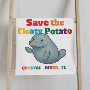 Puede incluir: Toalla blanca con un manatí de dibujos animados y el texto "Save the Floaty Potato" en colores arcoíris. Debajo del manatí está el texto "Crystal River, FL" en colores arcoíris. La toalla está colgada en un estante de madera.