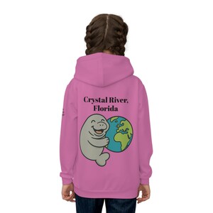 May include: Pink hooded sweatshirt with a cartoon manatee hugging a globe. The text "Crystal River, Florida" is above the graphic. The sweatshirt is a solid pink colour.
