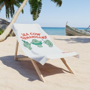 May include: A white beach chair with a canvas seat featuring the text "SEA COW SHENANIGANS" in red. The canvas displays cartoon manatees. The chair is made of light-coloured wood and is set on a sandy beach with a boat in the background.