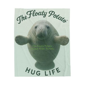 May include: A light green blanket with a manatee illustration. The text "The Floaty Potato" and "Hug Life" are displayed. The manatee is grey with outstretched arms. The text "The Floaty Potato Crystal River, Florida" is also included.