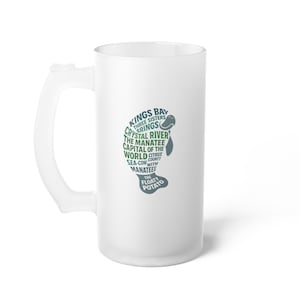 May include: Frosted white beer mug with a handle. The mug has a green and blue graphic of a manatee with text that reads "Kings Bay, Crystal River, The Manatee, Capital of the World, Sea-Cow with Manatees, The Floaty Potato."
