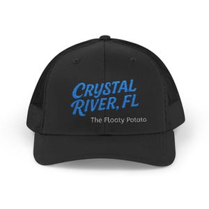 May include: Black trucker cap with a mesh back and a curved peak. The front displays the text "CRYSTAL RIVER, FL" in blue script, with "The Floaty Potato" underneath.