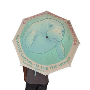 May include: A beige umbrella with a light blue manatee illustration. The umbrella has text that reads "Crystal River, Florida" and "Manatee Capital of the World." The text "The Floaty Potato" is also printed on the umbrella.