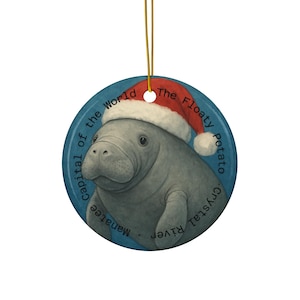 May include: A circular Christmas tree decoration depicting a grey manatee wearing a red Santa hat. The blue background has text that reads "Capital of the World Manatee Crystal River" and "The Floaty Potato."