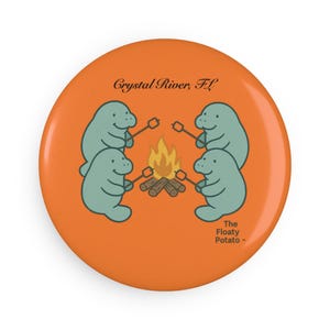 May include: An orange button featuring a cartoon of four light blue manatees toasting marshmallows over a campfire. The text "Crystal River, FL" and "The Floaty Potato" are also on the button.