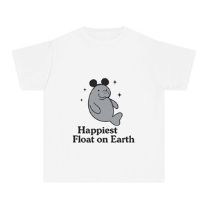 Puede incluir: Camiseta blanca con un manatí gris con orejas de ratón negras y el texto "Happiest Float on Earth". El diseño incluye estrellas. Un diseño divertido e informal.
