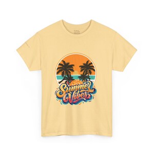 May include: A pale yellow t-shirt with a circular design showing palm trees, a sunset, and the words "Summer Vibes" in a colourful, stylised font. The shirt is made of a soft material.