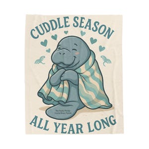 May include: A light beige blanket with a cartoon manatee wrapped in a striped blanket. The text "Cuddle Season All Year Long" is displayed in teal, with teal hearts and decorative elements. The text "The Floaty Potato Crystal River, Florida" is also included.