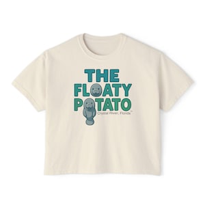 May include: A cream-coloured t-shirt with the text "THE FLOATY POTATO" in teal and green, featuring a cartoon manatee. Below the text is "Crystal River, Florida" in green. The shirt is a casual, short-sleeved style.