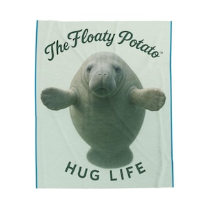 May include: A light blue blanket with a cartoon manatee design. The text "The Floaty Potato" is above the manatee, and "HUG LIFE" is below. The blanket has a light blue border. The manatee has outstretched arms.