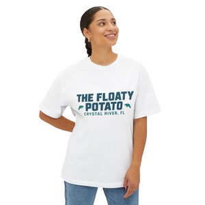 May include: White t-shirt featuring the teal text "THE FLOATY POTATO" and "CRYSTAL RIVER, FL". The design includes small teal manatee illustrations. The shirt has a loose fit and short sleeves.