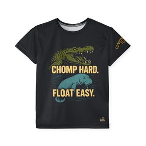 May include: Black t-shirt featuring a green alligator and a blue manatee graphic. The text "CHOMP HARD. FLOAT EASY." is printed below the alligator and manatee. The shirt also has the text "CRYSTA FLO" and "THE FLOATY POTATO" printed on it.