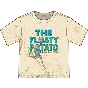 May include: A beige short-sleeve t-shirt with a distressed texture and a graphic print. The text reads THE FLOATY POTATO with two cartoon manatees integrated into the lettering. Below the text, it says Crystal River, Florida in small font.