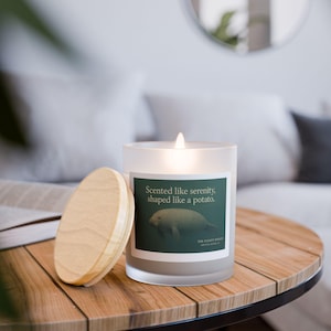 May include: A lit, frosted glass candle with a wooden lid on a wooden table. The candle has a label that reads "Scented like serenity, shaped like a potato." The candle's theme is relaxation and home fragrance.