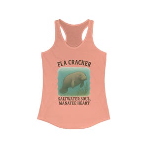 Può includere: Una canotta color corallo con una grafica di un lamantino che nuota sott'acqua. Il testo "FLA CRACKER" è sopra l'immagine e "SALTWATER SOUL, MANATEE HEART" è sotto.