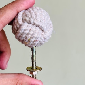 Może przedstawiać: Beżowy węzeł z liny przymocowany do metalowej śruby z podkładką i nakrętką. Węzeł z liny jest okrągły i ma fakturę. Metalowe elementy są w kolorze złotym.