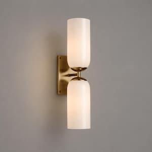 May include: A wall sconce featuring a rectangular brass backplate and two cylindrical, frosted white glass shades arranged vertically. The fixture includes a central brass arm connecting the two light sources, creating a balanced, linear design.