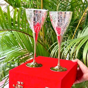 Pode incluir: Duas taças de champanhe ornamentadas, com taças prateadas e hastes douradas, estão sobre uma caixa de veludo vermelha. As taças têm um padrão floral detalhado. A caixa tem fechos e dobradiças douradas.