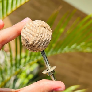 Puede incluir: Un pomo decorativo para armario con una esfera hecha de hilo natural bien enrollado. La bola de hilo está unida a un tornillo de metal con una arandela y una tuerca. El fondo es follaje verde borroso.