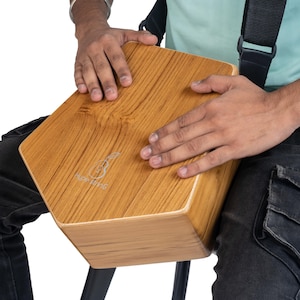 May include: A light brown wooden cajon drum with a wood grain pattern. The hexagonal drum has the "Hype Strings" logo. A black strap is visible. The drum is likely made of wood.