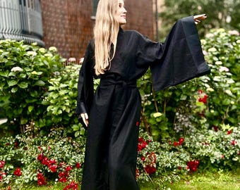 May include: A long, black kimono-style robe with wide sleeves and a matching belt. The robe has a simple design and is made of a flowing fabric. The person is standing in a garden with green grass and red flowers.
