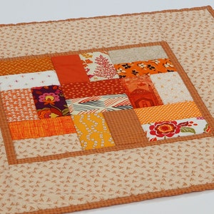 May include: A rectangular quilted fabric mat featuring a central patchwork panel of various orange, yellow, and red patterned squares. The border is a light beige fabric with a small repeating brown leaf print, finished with a thin orange checkered trim.