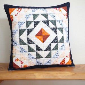 May include: A square throw pillow featuring a patchwork quilt design with a central orange and brown diamond motif. The pattern includes geometric triangles in dark green, white, and floral prints, all framed by a solid dark navy blue border.