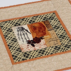 Peut inclure: Un tapis en tissu matelassé carré avec un panneau central illustrant un crâne, une main squelettique, une araignée et une rose sombre. Le texte "things that go bump in the night" est imprimé sur le panneau. Bordure à carreaux noirs et beiges.