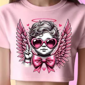Puede incluir: Top corto rosa pálido con un diseño de querubín. El querubín lleva gafas de sol en forma de corazón, alas rosas y un halo. Está haciendo el signo de la paz. Un gran lazo rosa y pequeños corazones completan el diseño.