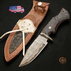 May include: A Damascus steel hunting knife with a dark wood handle and a leather sheath. The sheath is brown with a white and black accent and a leather strap closure. The knife blade has a unique pattern.