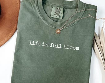 May include: An olive green t-shirt with the phrase "life in full bloom" printed in white. A gold necklace with a pendant rests on the shirt. The shirt is labeled "Comfort Colors" and is a size small.