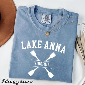 May include: A blue jean Comfort Colors t-shirt with the words "LAKE ANNA VIRGINIA" in white block letters. The design includes crossed oars. A gold necklace with a pendant is on the shirt.
