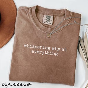 Puede incluir: Camiseta color espresso con la frase "whispering why at everything" en blanco. La camiseta tiene cuello redondo y una pequeña etiqueta que dice "Comfort Colors". Un collar plateado con un colgante está drapeado sobre la camiseta.