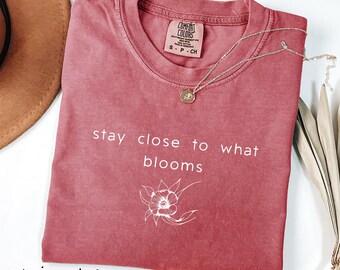 May include: Crimson t-shirt with the text "stay close to what blooms" and a floral illustration. A gold necklace with a pendant rests on the shirt. The shirt has a Comfort Colors tag.