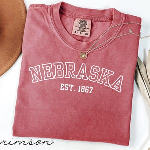 Op de afbeelding: Kleur kersrood T-shirt met het woord "NEBRASKA" in witte blokletters en "EST. 1867" eronder. Een gouden ketting met een hanger ligt op het shirt. Het shirt is opgevouwen en heeft een label met "Comfort Colors".