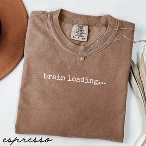 Peut inclure: T-shirt couleur expresso avec l'inscription blanche "brain loading...". Le t-shirt a un col rond et des manches courtes. Un collier argenté avec un pendentif est drapé sur le t-shirt. Le t-shirt est fait d'une matière douce et confortable.
